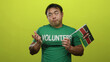 © Krakenimages.com - Asian man holding kenyan flag while wearing volunteer shirt, standing against vibrant yellow background, expressing confusion and uncertainty in isolated frame.