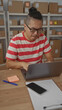 © Krakenimages.com - Man typing on laptop at wooden desk with parcel shelves in a building office, wearing red striped shirt and glasses; frustration small business.