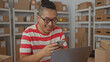 © Krakenimages.com - Man wearing glasses holding smartphone and typing with both hands beside laptop amid stacked parcel boxes on shelves in an office building, smiling; small business productivity.