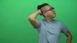 © Krakenimages.com - Young chinese man wearing glasses and light blue tshirt with hand behind head and squinting expression in green studio; thinking confusion.