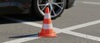 © olga_demina - Orange and white traffic cone standing on asphalt pavement with white lines, a car wheel and body visible in the background, symbolizing a driving lesson or test