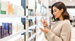 © Mariya Sorvacheva - Young woman shopping for cosmetics in a modern store, examining product packaging while surrounded by a variety of skincare and beauty items, showcasing a vibrant retail environment