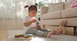 © sorrapongs - A two year old Asian girl sits barefoot on a soft gray rug, stacking ring toys near sheer curtains, a beige sofa, patterned pillows, and a plush teddy bear.