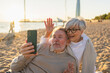 © Юлия Завалишина - Video call. Senior couple making selfie having video call on beach outdoor. Mature old grandmother grandfather talking speaking online. Older generation modern tech usage. Virtual meeting online chat