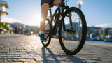 Woman cycling in a city during late afternoon, close-up on legs pedaling and wheels turning, sunlight casting long shadows, textured pavement and urban background slightly blurred,