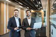 © Liubomir - Two smiling adult businessmen wearing suits standing in a corporate office corridor, one holding a clipboard, the other a laptop, representing collaboration and professional success