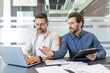 © Liubomir - Two male business colleagues working together, discussing ideas and planning strategies while reviewing data on a laptop and documents during a meeting in a contemporary workspace