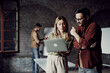 © Novak - Two young entrepreneurs in casual clothes looking at a laptop screen and discussing a project, while a colleague works in the background of a modern creative office.
