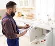 © New Africa - Plumber in uniform with clipboard writing in kitchen