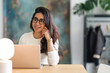 © Studio Marmellata - A young woman with glasses smiles while working on her laptop at a desk, with a lamp and artwork in the background.