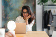 © Studio Marmellata - A woman wearing glasses smiles while looking at her laptop. She is sitting at a wooden desk with a lamp and a phone.