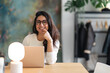 © Studio Marmellata - A smiling woman with glasses sits at a desk with a laptop and a lamp, working in a modern office space.