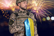 © New Africa - Soldier holding flag of Ukraine near his heart against blurred background with fireworks. National holiday greeting card or poster design