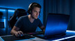 © SilhouetteARTs - Focused young man intensely gaming on a laptop with headphones in a dark room illuminated by blue light, concentrated on online competition.