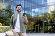 © Liubomir - Happy man standing outside a modern office building, smiling while using his smartphone, representing urban lifestyle, connectivity, and communication in a business environment
