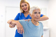 © Graphicroyalty - professional female physiotherapist in blue scrubs providing manual guidance to a senior woman performing arm and shoulder rotation exercises in a bright, modern rehabilitation clinic ward.
