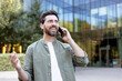 © Liubomir - Smiling man standing outdoors, engaging in a pleasant phone conversation while gesturing with his hand, with a modern glass building and green trees blurring in the background
