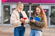 © Iryna - Two young women stand outside a building while looking at their phones and holding books