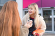 © Iryna - Two young women talk and laugh outside a building while holding books and a phone