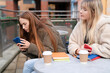 © Iryna - Friends sit outside at a cafe and use their phones while enjoying warm drinks on a cool afternoon