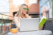 © Iryna - Woman reading a book while sitting at a table with a laptop and coffee in an outdoor cafe