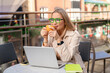 © Iryna - Woman sits outside with laptop while enjoying coffee on a sunny day in a busy plaza