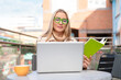 © Iryna - Person reads book and works on laptop at outdoor cafe during the day