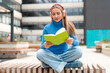 © Iryna - Reading a book while wearing headphones in an outdoor space near a modern building