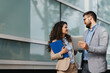 © Novak - Businessman and businesswoman standing in front of a modern office building, discussing a project using a digital tablet and paper documents