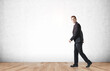 © ImageFlow - Confident businessman walking in formal suit on wooden floor with white wall background, representing business progress and creative concept