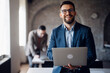 © Novak - Smiling businessman holding a laptop and looking at the camera with a colleague in the background.