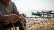 © BerkahStock - Elderly fisherman meticulously repairing a fishing net by the sea with a boat in the background.