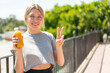 © luismolinero - Young blonde woman holding an orange juice at outdoors smiling and showing victory sign