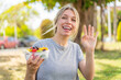 © luismolinero - Young blonde woman holding a bowl of fruit at outdoors saluting with hand with happy expression