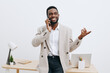 © SHOTPRIME STUDIO - Confident smiling African American man in beige blazer talking on mobile phone while standing at modern office desk, using laptop and stationery, studio shot. Business communication concept