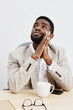 © SHOTPRIME STUDIO - Thoughtful young man with dark skin, short curly hair, wearing beige blazer and white shirt, sitting at desk with coffee mug, smartphone, and glasses, looking upward with hands clasped, studio shot