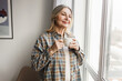 © shurkin_son - Adorable caucasian smiling senior lady in plaid shirt enjoying her herbal tea standing in cozy room next to big window and admiring first snow and winter coming looking through at city street