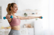 © Prostock-studio - Covid-19 quarantine, home gym, body care and new normal. Smiling millennial blonde woman in sports uniform doing exercises with dumbbells for arms in minimalist living room interior, copy space