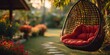 © horizon - Hanging rattan egg chair with red cushions suspended in a beautiful green garden, backlit by golden sunlight for a dreamy backyard scene.