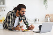 © Prostock-studio - Irritated middle-eastern guy having snack while working from home during coronavirus pandemic, young arab man eating healthy food fresh salad, holding phone and looking at laptop screen, copy space