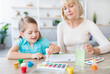 © Prostock-studio - Loving old grandmother drawing picture together with small granddaughter at home. Caring granny involved in playful funny learning activity with little grandchild using watercolor painting