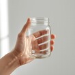 © Shamim - Hand holding empty clear glass jar against white background minimalist concept