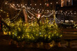 © Viktoria - Festive winter night scene in Vilnius Old Town city park. Small potted fir trees wrapped in fabric glow with warm yellow fairy lights, arranged in a row on a paved square. Overhead string lights hang