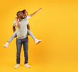 © Prostock-studio - Discounts for family and kids. Excited black man giving piggyback ride for little daughter, cute girl pointing aside at empty space over yellow background, studio shot