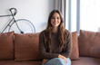 © luciano - Portrait of a smiling young woman with long brown hair sitting on sofa in a cozy modern living room looking at camera. Natural light, warm tones. Concept of happiness, confidence, and everyday life