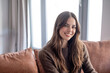© luciano - Portrait of a smiling young woman with long brown hair sitting on sofa in a cozy modern living room looking at camera. Natural light, warm tones. Concept of happiness, confidence, and everyday life