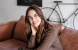 © luciano - Portrait of a smiling young woman with long brown hair sitting on sofa in a cozy modern living room looking at camera. Natural light, warm tones. Concept of happiness, confidence, and everyday life