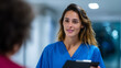 © Alexander - Hyperrealistic commercial stock photo: Latina nurse (25-35) in scrubs communicating with one adult patient in modern hospital corridor, pointing direction, clipboard, sharp eyes BO