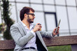 © Mediteraneo - Young businessman sitting on a bench in front of a modern office building, drinking coffee and using a tablet.