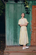 © SHOTPRIME STUDIO - Elegant woman in a pale yellow dress leans against a rustic green door, posing thoughtfully in an outdoor setting with soft light, calm mood, and simple surroundings